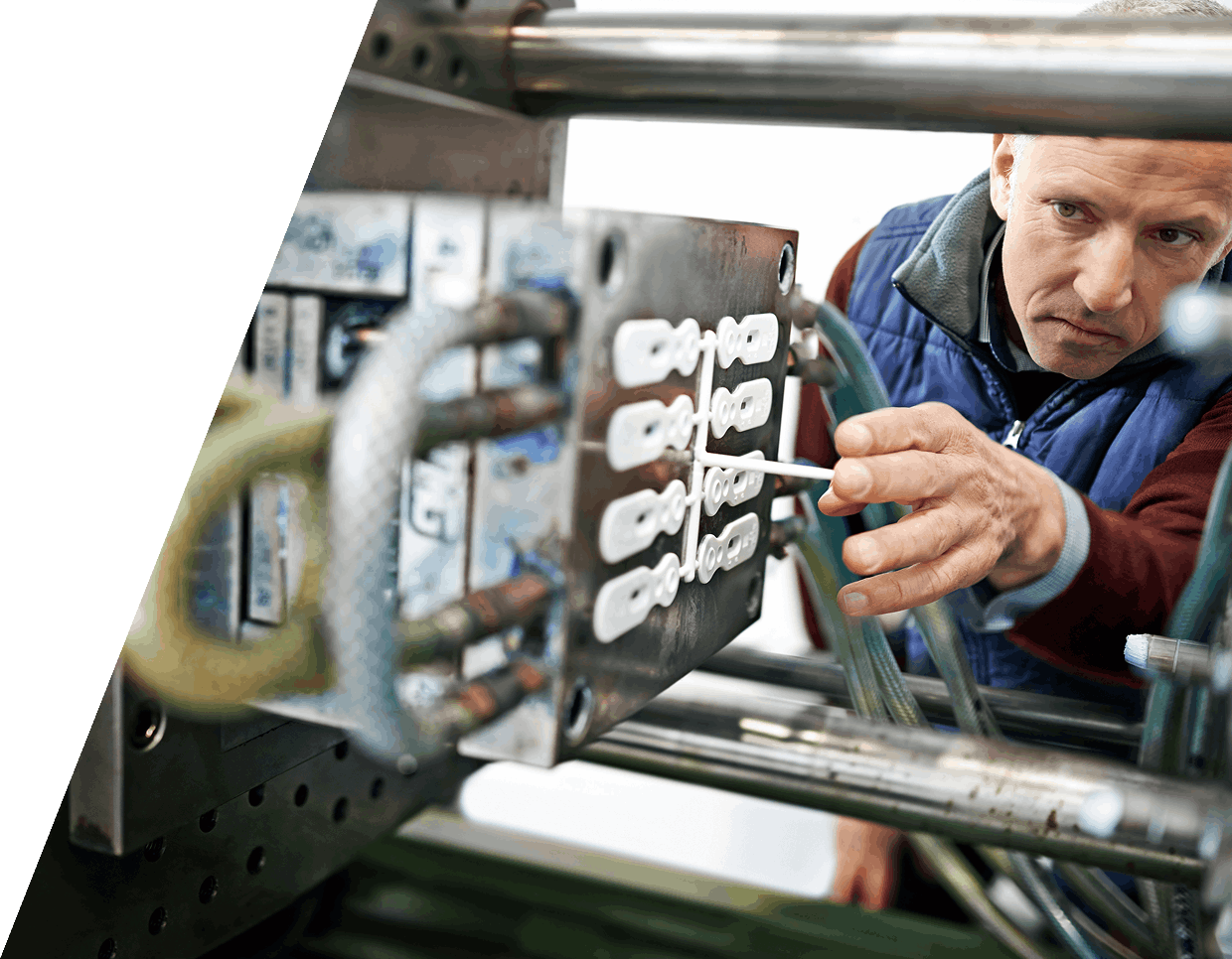 jpeg-optimizer_GettyImages-1371069614 Man inspecting a mold for plastic manufacturing inside a factory, wearing a blue jacket and holding a tool.