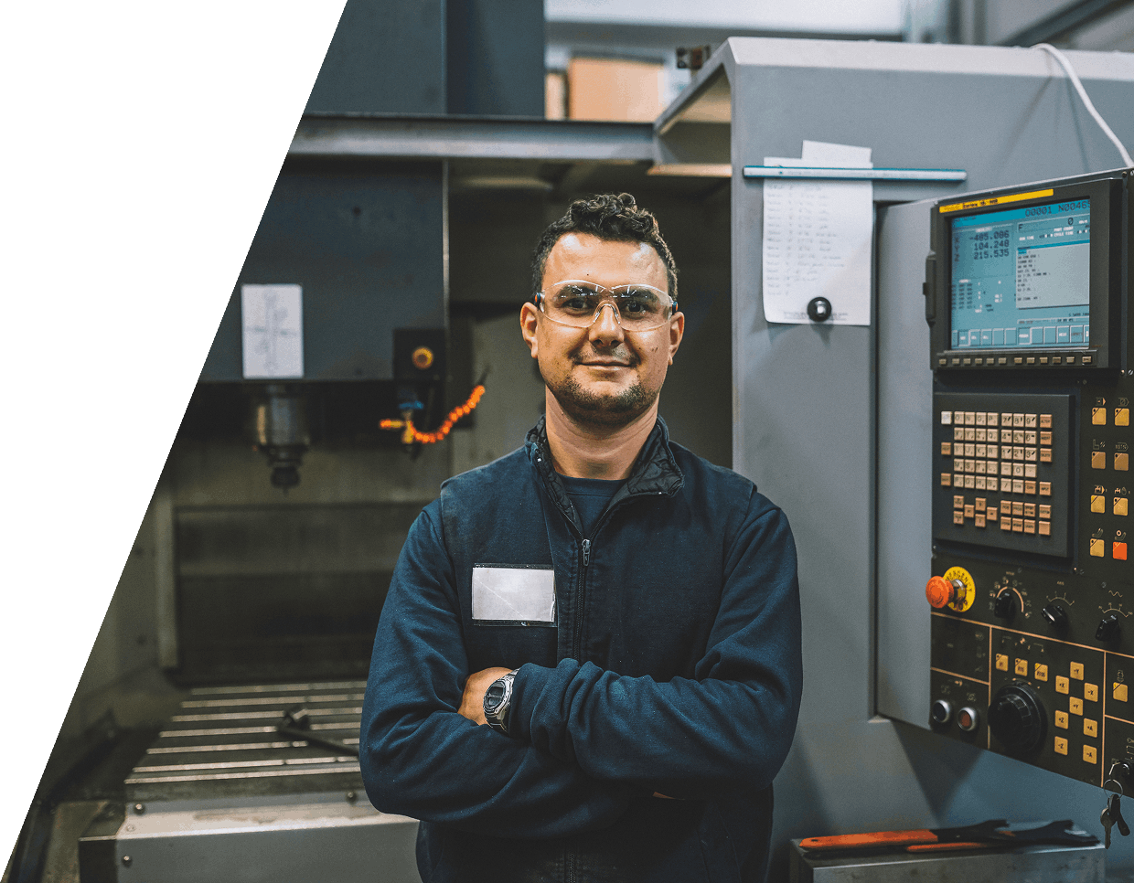 jpeg-optimizer_GettyImages-1371069614 Man in safety glasses and blue coverall stands with arms crossed in front of industrial machinery, next to a control panel with buttons and display.