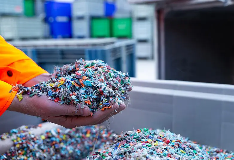 jpeg-optimizer_GettyImages-2065999665 Hands holding a mixture of colorful shredded plastic pieces above a container, with industrial bins in the background.